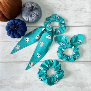 Puede incluir: Gomas para el pelo y lazos para el pelo de tela turquesa con un estampado de calaveras blancas. Las gomas para el pelo tienen varios estilos, incluido uno en forma de lazo. En el fondo hay calabazas decorativas marrones, azules y grises. Ideal para Halloween.