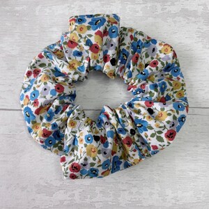 Puede incluir: Una scrunchie floral con fondo blanco y un estampado de flores azules, rojas y amarillas. La tela est&aacute; fruncida, creando un efecto de volantes. Este accesorio para el cabello es una adici&oacute;n elegante a cualquier peinado.