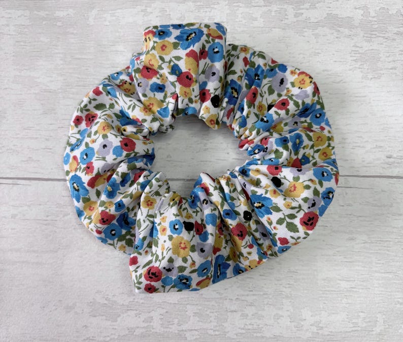 Puede incluir: Una scrunchie floral con fondo blanco y un estampado de flores azules, rojas y amarillas. La tela est&aacute; fruncida, creando un efecto de volantes. Este accesorio para el cabello es una adici&oacute;n elegante a cualquier peinado.