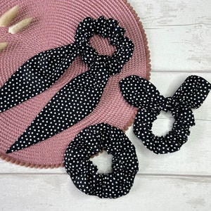 Puede incluir: Cuatro coleteros y cintas para el pelo negros y blancos con estampado de corazones están colocados sobre un salvamanteles rosa. Los coleteros son redondos, mientras que las cintas tienen extremos largos y puntiagudos. Fondo de madera clara.