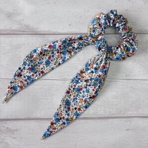 Puede incluir: Una goma para el pelo floral con lazos largos y puntiagudos de tela. La goma y los lazos presentan un estampado de peque&ntilde;as flores azules, rojas y amarillas sobre fondo blanco. Accesorio para el cabello.