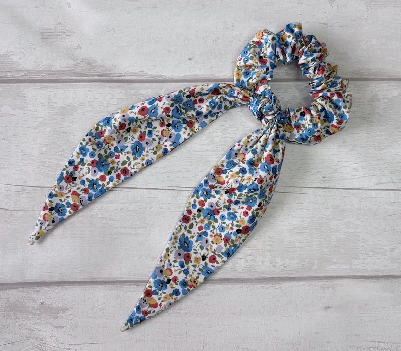 Puede incluir: Una goma para el pelo floral con lazos largos y puntiagudos de tela. La goma y los lazos presentan un estampado de peque&ntilde;as flores azules, rojas y amarillas sobre fondo blanco. Accesorio para el cabello.