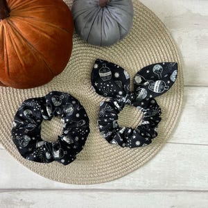 May include: Two black and white hair scrunchies with a potion bottle print. The scrunchies are on a woven straw-like surface.