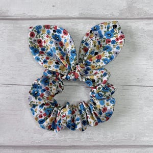 Puede incluir: Una scrunchie floral con detalles de orejas de conejo. La tela presenta un patr&oacute;n de peque&ntilde;as flores azules, rojas y amarillas sobre un fondo blanco. Este accesorio para el cabello est&aacute; dise&ntilde;ado para sujetar el cabello y a&ntilde;ade un elemento decorativo.