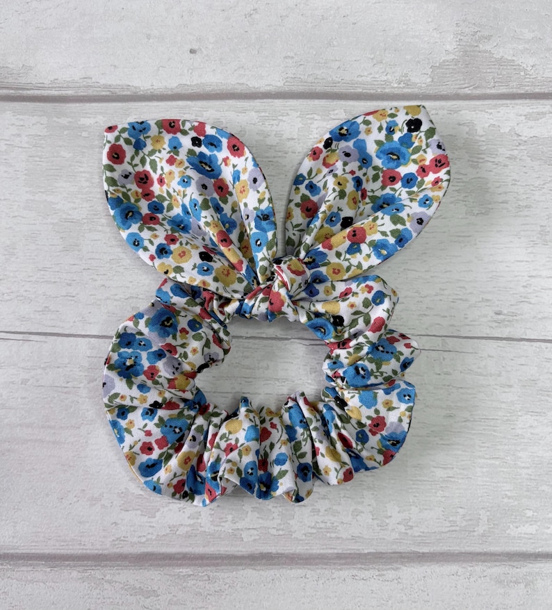 Puede incluir: Una scrunchie floral con detalles de orejas de conejo. La tela presenta un patr&oacute;n de peque&ntilde;as flores azules, rojas y amarillas sobre un fondo blanco. Este accesorio para el cabello est&aacute; dise&ntilde;ado para sujetar el cabello y a&ntilde;ade un elemento decorativo.