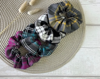 May include: Four plaid scrunchies in various colors: magenta, teal, black and white, and gray with yellow accents. The scrunchies are arranged on a neutral-toned, circular woven mat, with dried decorative grass.
