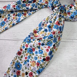 Puede incluir: Una goma para el pelo floral con lazos de tela. La tela presenta un fondo blanco con un estampado de peque&ntilde;as flores azules, rojas, amarillas y moradas. La goma para el pelo est&aacute; atada con un lazo.