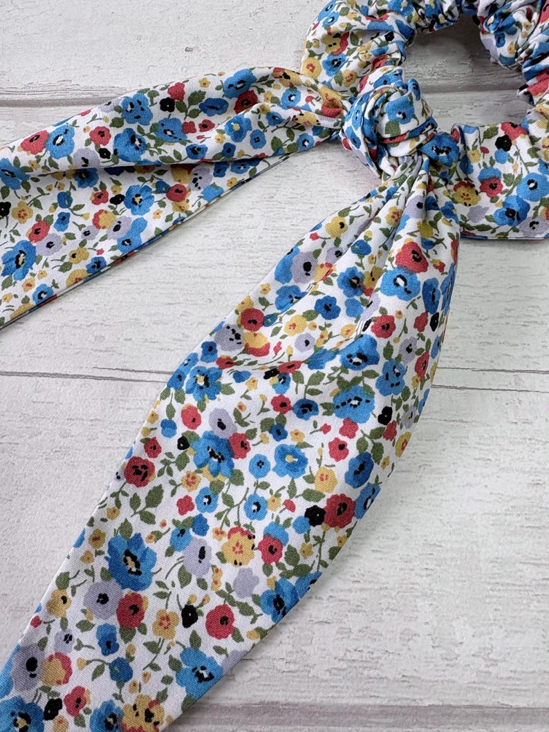 Puede incluir: Una goma para el pelo floral con lazos de tela. La tela presenta un fondo blanco con un estampado de peque&ntilde;as flores azules, rojas, amarillas y moradas. La goma para el pelo est&aacute; atada con un lazo.