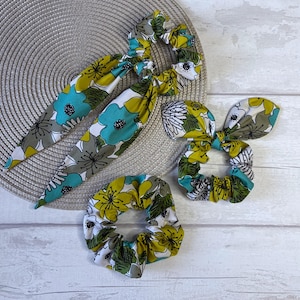 Puede incluir: Un conjunto de cuatro accesorios para el cabello con motivos florales, que incluye dos diademas y dos coleteros. La tela presenta un estampado de grandes flores turquesas, amarillas y blancas con detalles grises. Dispuestos sobre una superficie texturizada de tono neutro.