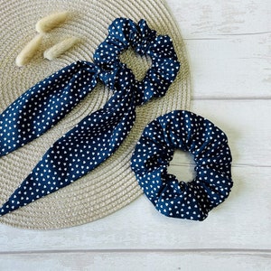 May include: Two hair scrunchies with a white heart pattern on a dark blue background. One scrunchie is a long, tied bow and the other is a simple circle.