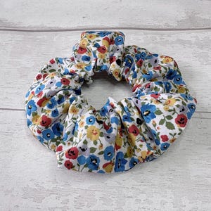 Puede incluir: Una scrunchie floral con fondo blanco adornada con peque&ntilde;as flores rojas, azules y amarillas. La tela est&aacute; fruncida y el&aacute;stica, formando una forma circular. Este accesorio para el cabello es adecuado para varios peinados.