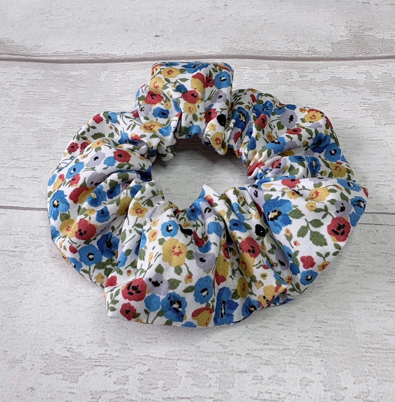 Puede incluir: Una scrunchie floral con fondo blanco adornada con peque&ntilde;as flores rojas, azules y amarillas. La tela est&aacute; fruncida y el&aacute;stica, formando una forma circular. Este accesorio para el cabello es adecuado para varios peinados.