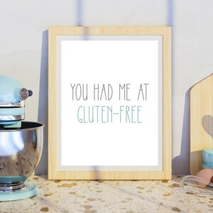 Pu&ograve; includere: Una stampa incorniciata con il testo "You had me at gluten-free" in lettere nere e turchese su sfondo bianco. La cornice &egrave; in legno chiaro e la stampa &egrave; appoggiata su un piano di lavoro marrone chiaro.