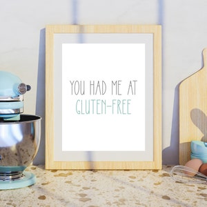 Pu&ograve; includere: Una stampa incorniciata con il testo "You had me at gluten-free" in lettere nere e turchese su sfondo bianco. La cornice &egrave; in legno chiaro e si trova su un piano di lavoro marrone chiaro.
