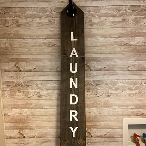 以下が含まれることがあります： 「LAUNDRY」の文字が白で縦に書かれた、濃い茶色の木製ランドリーサイン。上部には黒い金属パイプが取り付けられ、下部には「Drop Drawers Here!」の文字があります。明るい茶色の木製パネルの壁に取り付けられています。