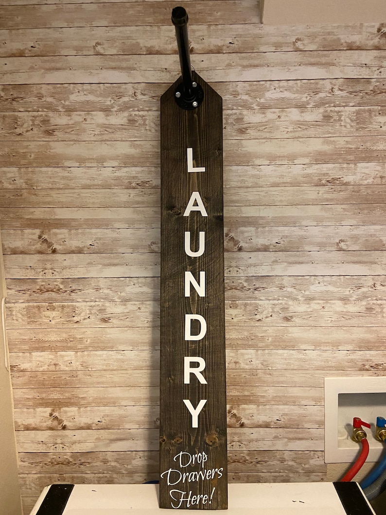 以下が含まれることがあります： 「LAUNDRY」の文字が白で縦に書かれた、濃い茶色の木製ランドリーサイン。上部には黒い金属パイプが取り付けられ、下部には「Drop Drawers Here!」の文字があります。明るい茶色の木製パネルの壁に取り付けられています。