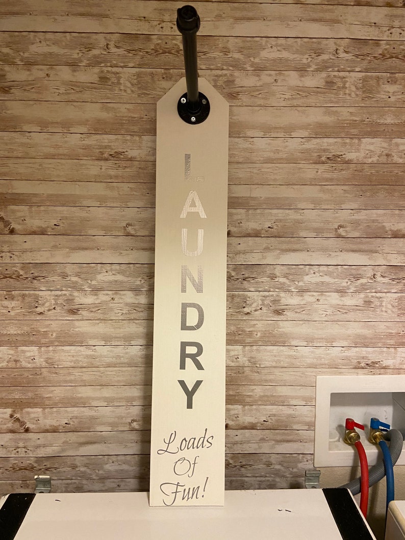 以下が含まれることがあります： 「LAUNDRY」の文字が大きくグレーで書かれた、長方形のオフホワイトのランドリーサイン。「Loads Of Fun!」のフレーズが筆記体で書かれています。黒いパイプとフランジで木製のパネルの壁に取り付けられています。洗濯室用です。