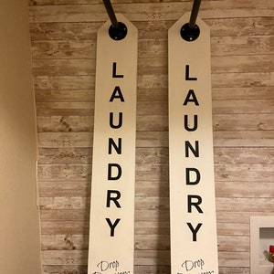 以下が含まれることがあります： 「LAUNDRY」と縦書きで書かれた黒い文字と、その下に「Drop Drawers Here!」と書かれた2つの白い木製のランドリーサイン。各サインの上部には黒い金属パイプが付いています。