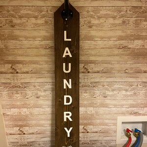 以下が含まれることがあります： 「LAUNDRY」と「Drop Drawers Here!」と書かれた白い文字が入った、濃い色の木製ランドリーサイン。上部には黒い金属パイプ継手が付いており、吊り下げることができます。木製のパネルが貼られた壁に取り付けられています。