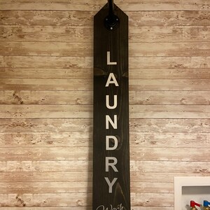 以下が含まれることがあります： 「LAUNDRY」と縦書きで書かれ、下部に「Wash Fold Repeat!」と書かれた白い文字が入った、濃い色の木製のランドリーサイン。黒い金属パイプが看板の上部から伸びています。看板は、明るい茶色の木製パネルの壁に取り付けられています。