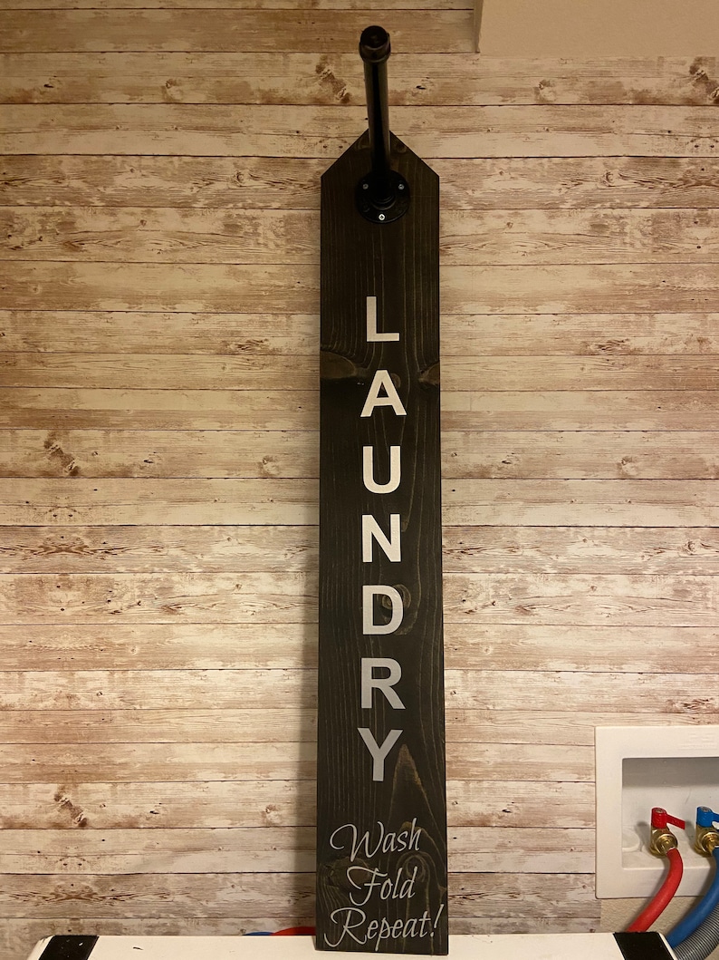 以下が含まれることがあります： 「LAUNDRY」と縦書きで書かれ、下部に「Wash Fold Repeat!」と書かれた白い文字が入った、濃い色の木製のランドリーサイン。黒い金属パイプが看板の上部から伸びています。看板は、明るい茶色の木製パネルの壁に取り付けられています。