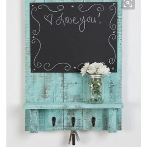 May include: A teal blue wooden key holder with a chalkboard sign that says "Love you!" and a small glass jar with white flowers. The key holder has four hooks for hanging keys.