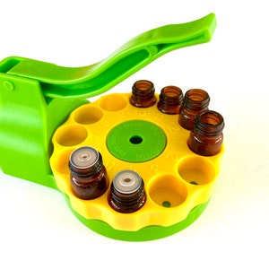 Op de afbeelding: Groene en gele plastic organizer voor essentiële olieflessen met een hendel om de flessen te openen en te sluiten. De organizer heeft 12 vakjes voor flessen en een genummerde wijzerplaat om het gebruik bij te houden.