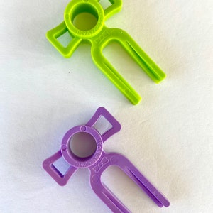 May include: Two plastic bottle openers, one green and one purple, shaped like a person with a bow tie and a fork-like opening. The text "My Aroma Pal" is printed on both openers.