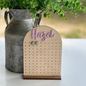 May include: A wooden earring display stand with the name "Hazel" in pink script. The stand has numerous small holes for holding earrings and a dark brown base. Two stud earrings are displayed.