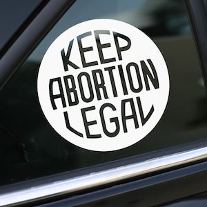 May include: A white circle sticker with black text that reads "Keep Abortion Legal" on a black car window.
