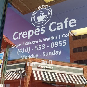 May include: A white sign on a glass door advertises "Irina's Crepes Cafe" with the phone number (410) - 553 - 0955. The sign also lists the cafe's hours: Monday - Sunday, 8am - 3pm. The cafe serves crepes, chicken and waffles, and coffee.