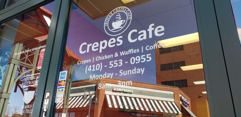 May include: A white sign on a glass door advertises "Irina's Crepes Cafe" with the phone number (410) - 553 - 0955. The sign also lists the cafe's hours: Monday - Sunday, 8am - 3pm. The cafe serves crepes, chicken and waffles, and coffee.