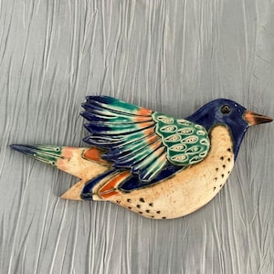 May include: A ceramic bird sculpture with a blue head, wings of teal, orange, and blue, and a cream-coloured body. The bird has a dark beak and is decorated with small black dots.