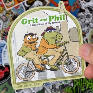 May include: A sticker featuring two cartoon characters riding a tandem bicycle. The sticker reads "Grit and Phil" and "A Little Book of Big Jawns." The sticker has a green border and the words "ROSE BEAR ARTS" at the bottom.