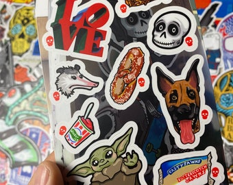 May include: A sheet of stickers featuring various designs, including a red "LOVE" sign, a pretzel, a skull, a dog, a possum, a green alien character, a red and white cup with a straw, and a brown and blue package with the text "Tasty Jawns Butterscotch Jawns".