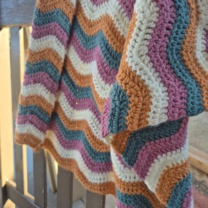 May include: A colorful, handmade crochet blanket with a chevron pattern. The blanket features stripes of white, orange, teal, and pink. The blanket is draped over a wooden railing, showcasing the texture and design.
