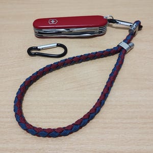 May include: A red Swiss Army knife with a silver carabiner and a navy blue and burgundy braided lanyard. The knife is closed and features a white cross emblem. The lanyard is looped.