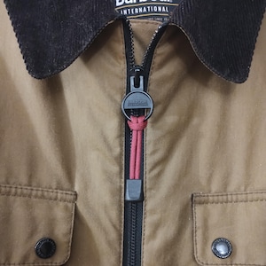 May include: A brown Barbour International jacket with a black zip and a black corduroy collar. The zip has a black pull with the Barbour International logo.