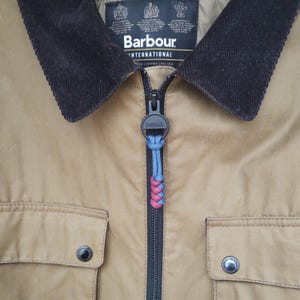 May include: Close-up of a brown Barbour International jacket with a black corduroy collar and a black zip. The zip pull has a black Barbour logo and is decorated with a braided cord in blue, red, and pink.