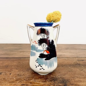May include: A small white ceramic vase with a blue rim and gold handles. The vase has a hand-painted design of a house and trees in a winter landscape. A yellow flower is in the vase.