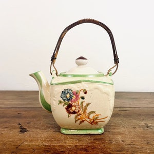 May include: A small, white ceramic teapot with a green and floral design. The teapot has a brown bamboo handle and a lid with a small knob.