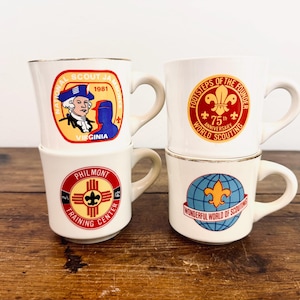 May include: Four white ceramic mugs with gold rims and various colorful designs. One mug features a historical figure and the text "National Scout Jamboree 1981 Virginia." Other mugs display scouting emblems and text, including "Philmont Training Center."