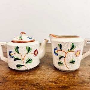 May include: A vintage white teapot and creamer set with floral designs in shades of blue, pink, green, and orange. The teapot has a lid and a handle, while the creamer has a spout and a handle.