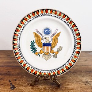 May include: A vintage decorative plate featuring the Great Seal of the United States. The plate has a white background with a colorful border of red, yellow, and black. The seal includes an eagle, stars, and the motto "E Pluribus Unum."