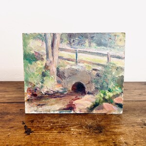 May include: An oil painting depicts a serene landscape with a small stone bridge over a stream. The artwork features a wooden fence, lush greenery, and a tree trunk, all rendered in soft, impressionistic brushstrokes. The color palette includes greens, browns, and blues.