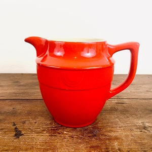 May include: A bright orange ceramic pitcher with a white rim and a decorative design. The pitcher has a rounded body and a handle.