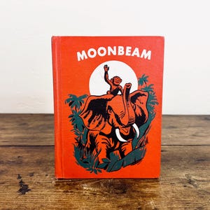 May include: A vintage children's book titled "MOONBEAM" with a vibrant orange cover. The cover features a cartoon illustration of an elephant with a monkey on its back, set against a white circle and green foliage. The book is displayed on a wooden surface.