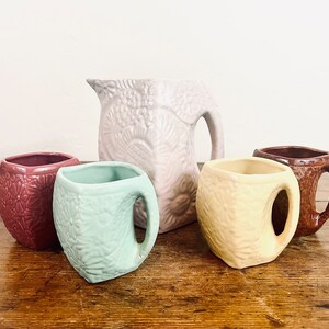 May include: Set of four ceramic mugs and a pitcher with embossed floral designs. The mugs are in shades of brown, green, and yellow, while the pitcher is a light purple.