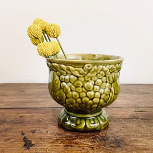 May include: A green ceramic pedestal vase with a textured, bubble-like pattern. The vase holds several yellow, spherical flowers. The vase is on a wooden surface, and the background is a plain white wall.