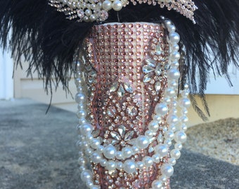 May include: A rose gold vase decorated with rhinestones and pearls. The vase is topped with a black feather boa and a rhinestone headband.